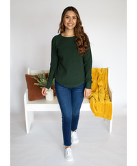 Woman wearing a green long sleeve and blue jeans standing in front of a decorated bench on a white background