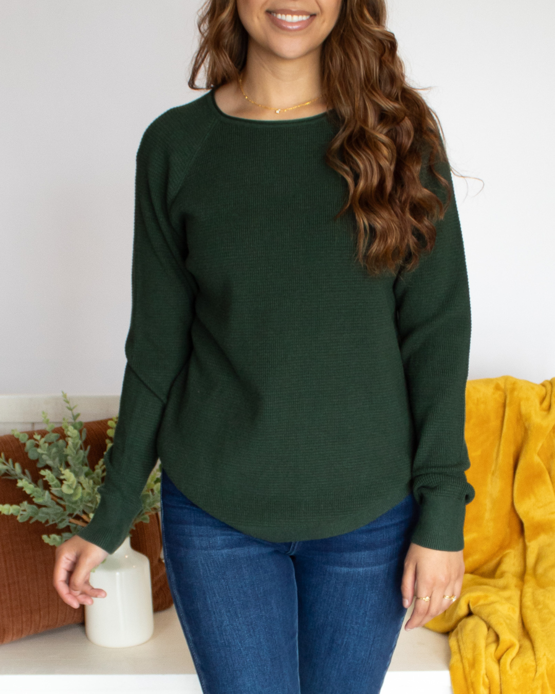 Close Up of a woman wearing a green long sleeve and blue jeans standing in a room with a white wall and decorated bench