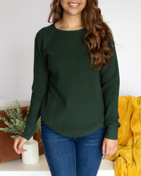 Close Up of a woman wearing a green long sleeve and blue jeans standing in a room with a white wall and decorated bench