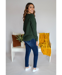 Woman wearing a green long sleeve and blue jeans standing in an angle in front of a white bench with decorative items.