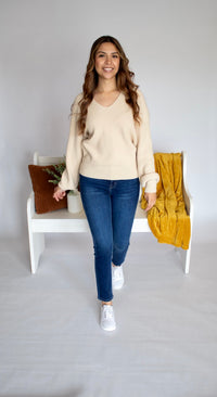 Woman wearing a beige sweater and blue jeans standing in a minimalistic room.
