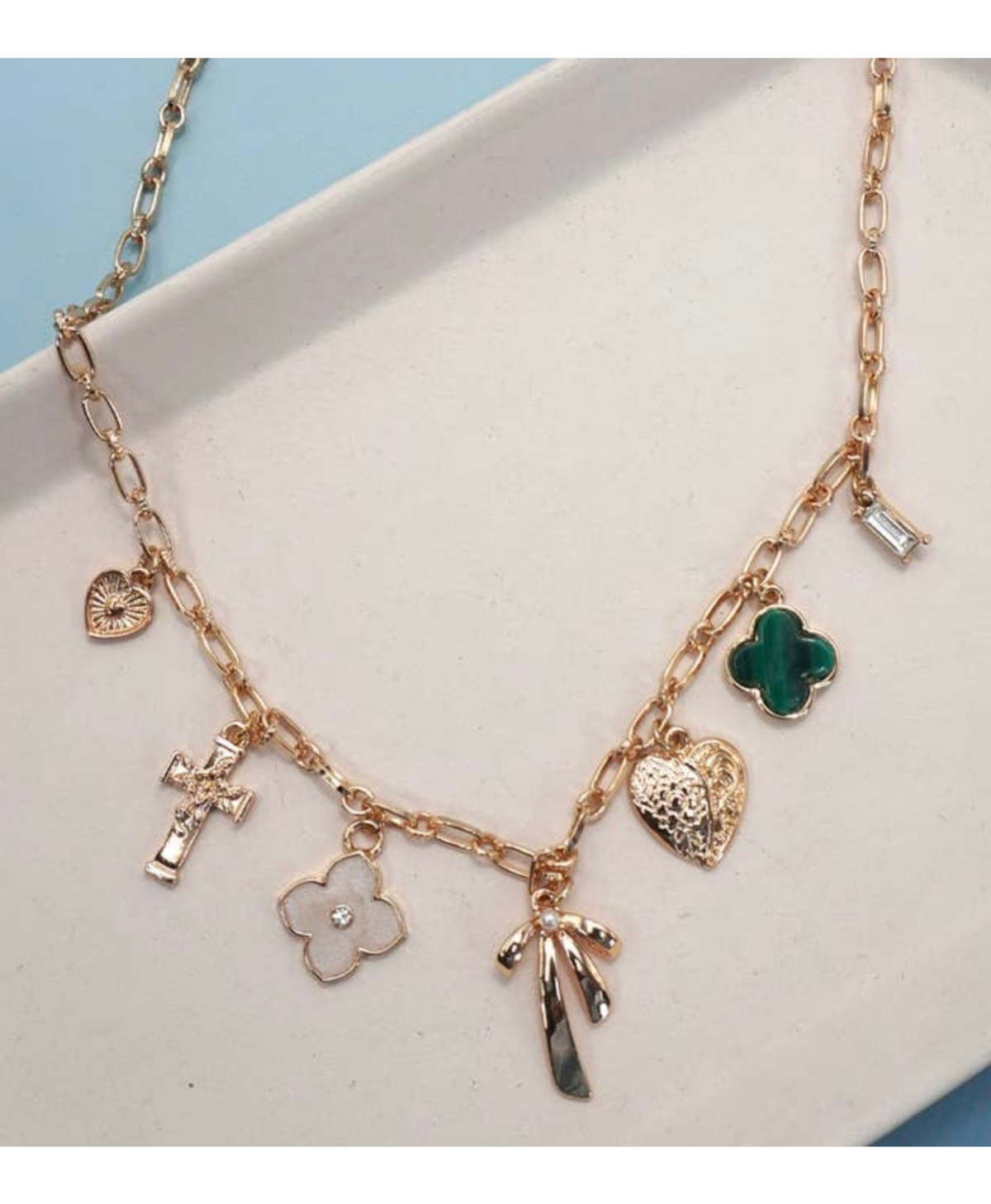 Gold necklace with various charms such as a heart, cross, floral design, bow, on a white surface with a light blue background