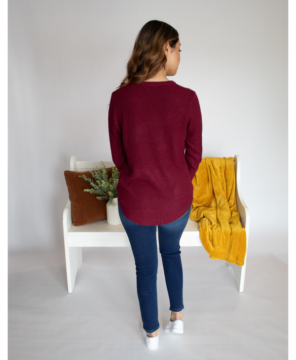 Back of woman wearing a maroon long sleeve and blue jeans standing in a minimalistic room with a white bench and yellow blanket.
