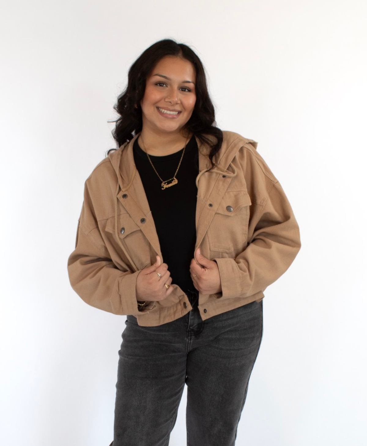 Woman wearing a cropped beige jacket on with a black shirt and black jeans on a white background
