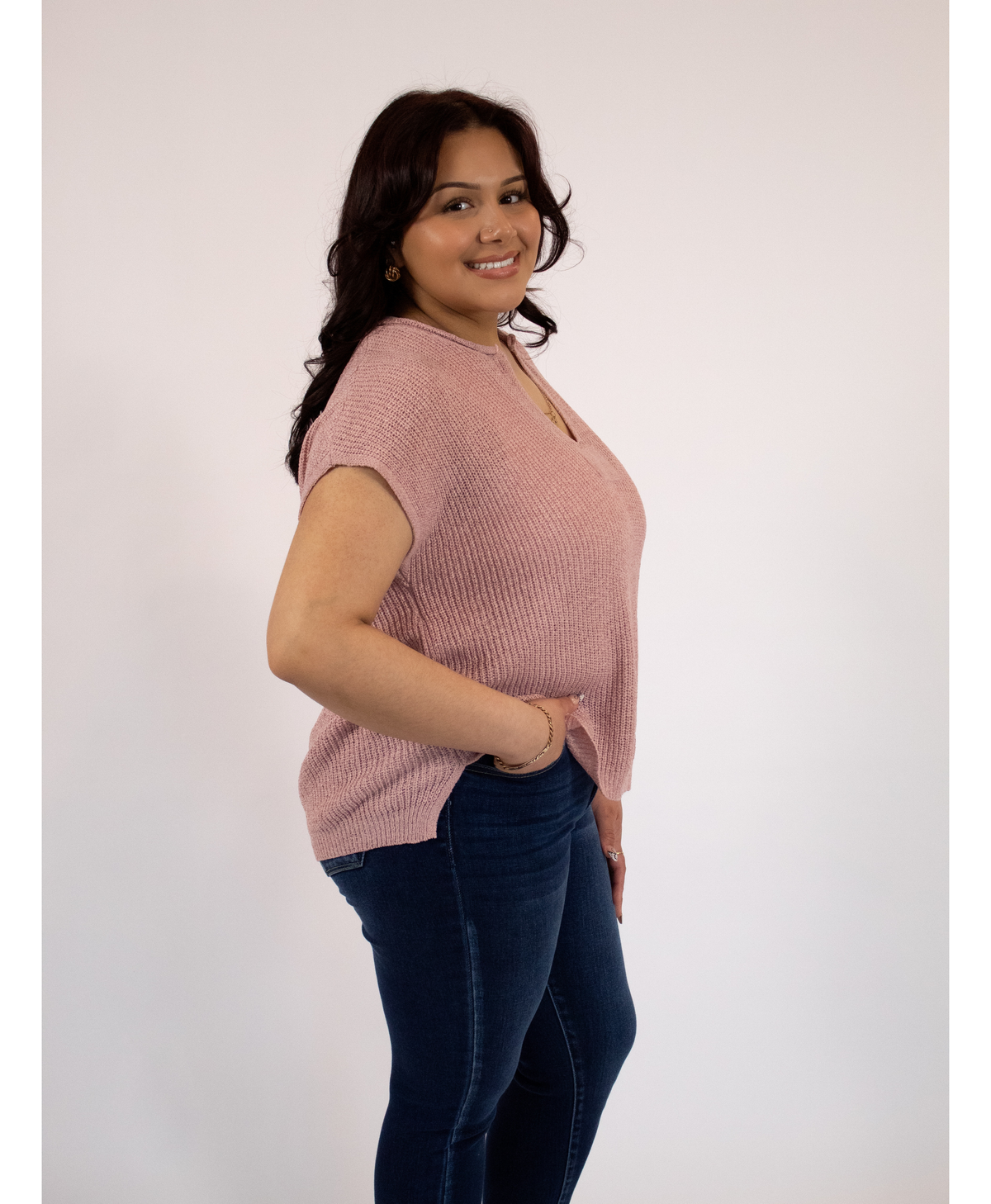 Side view of a woman wearing a pink split neckline top and blue jeans against a white background