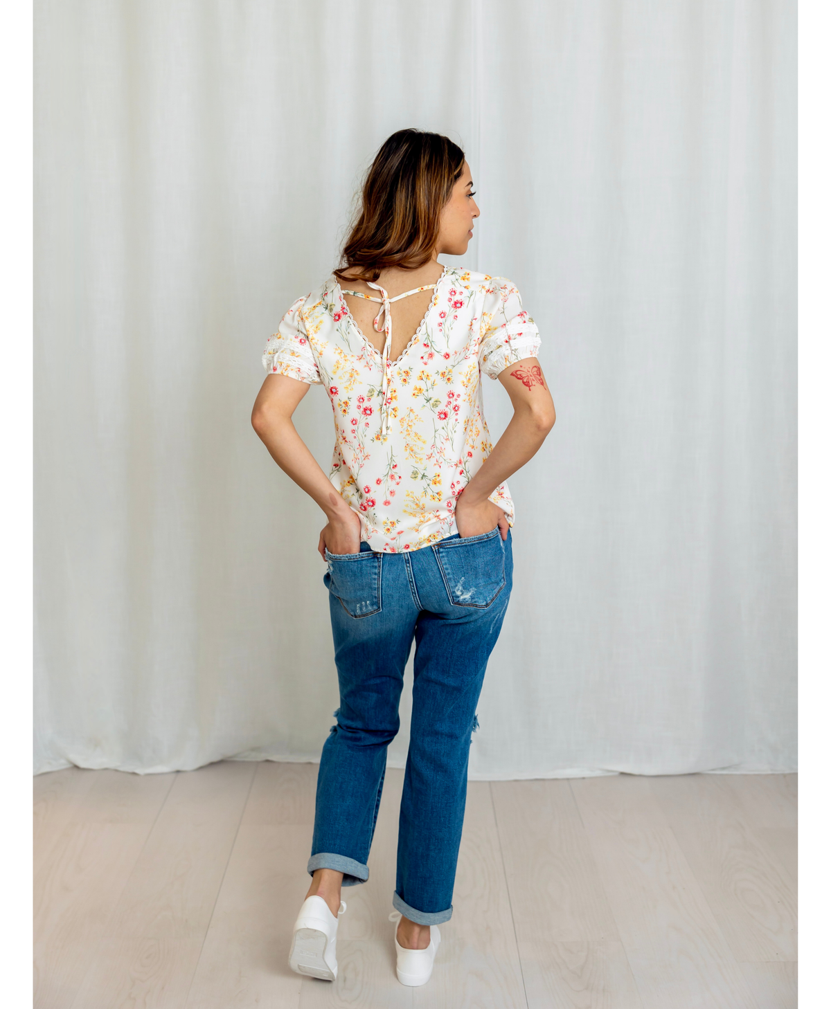 Back of women wearing a floral blouse with a v neck and bow in the back with a blue jeans with a plain background