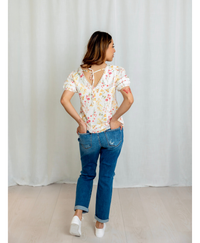 Back of women wearing a floral blouse with a v neck and bow in the back with a blue jeans with a plain background