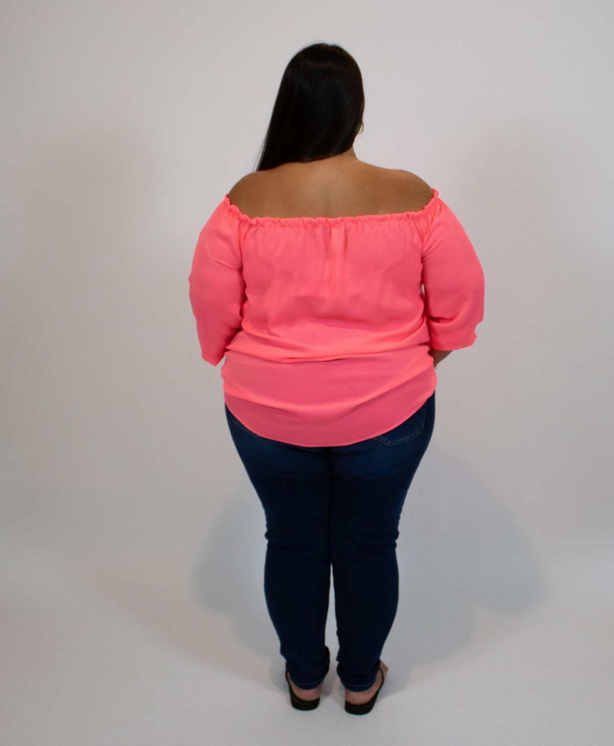 Person wearing a neon pink off-shoulder top and dark blue jeans on a plain background