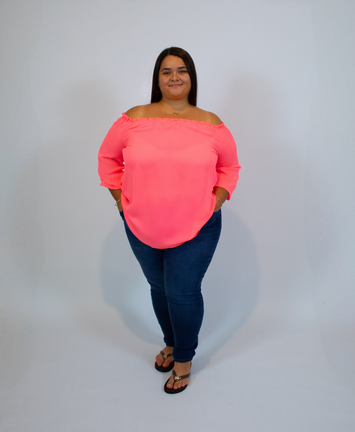 Woman wearing a neon pink off-shoulder top and dark blue jeans on a white background