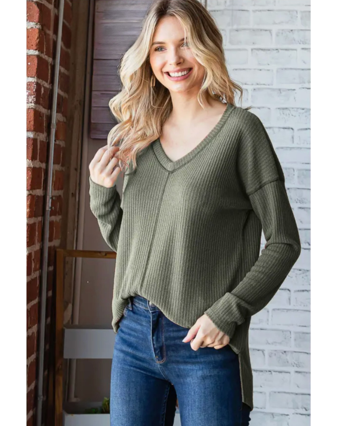 Woman wearing a green sweater and blue jeans standing against a brick wall.