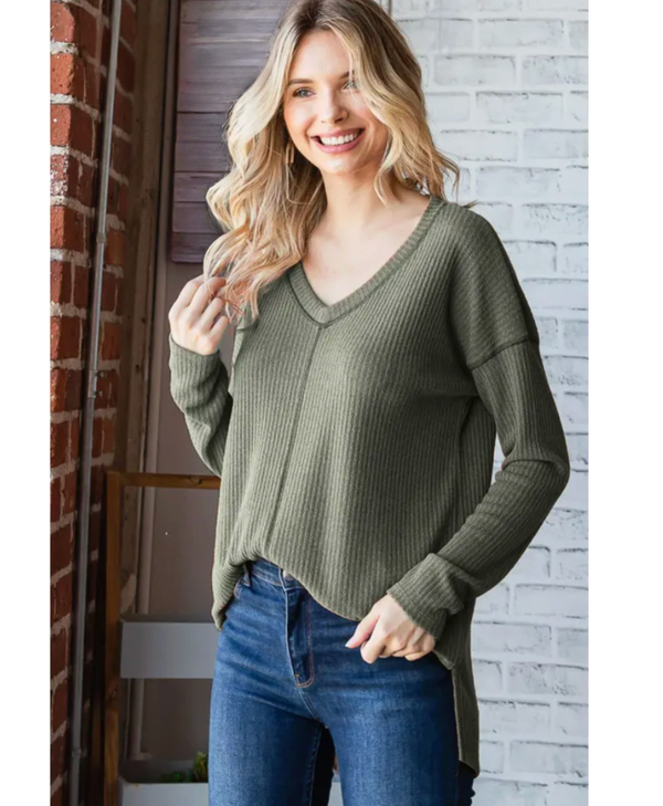 Woman wearing a green sweater and blue jeans standing against a brick wall.